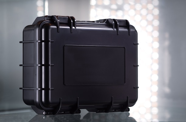 A sturdy black protective case with a rectangular shape and reinforced edges, set against a reflective surface. The case features multiple latches and a secure handle for easy transport, with a simple industrial design.