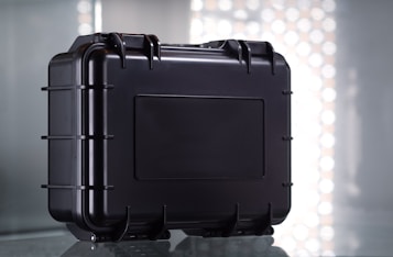 A sturdy black protective case with a rectangular shape and reinforced edges, set against a reflective surface. The case features multiple latches and a secure handle for easy transport, with a simple industrial design.