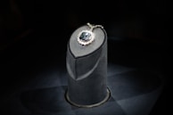 A luxurious gemstone necklace is displayed on a dark, elegant pedestal. The central gemstone is a deep, captivating blue, surrounded by smaller sparkling diamonds that enhance its brilliance. The spotlight enhances the sparkling effect, drawing attention to the intricate details and design.
