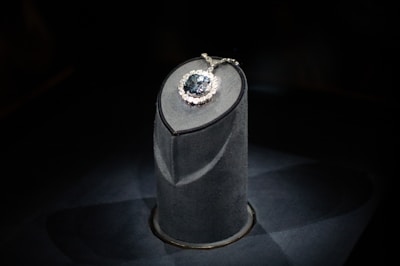 A luxurious gemstone necklace is displayed on a dark, elegant pedestal. The central gemstone is a deep, captivating blue, surrounded by smaller sparkling diamonds that enhance its brilliance. The spotlight enhances the sparkling effect, drawing attention to the intricate details and design.