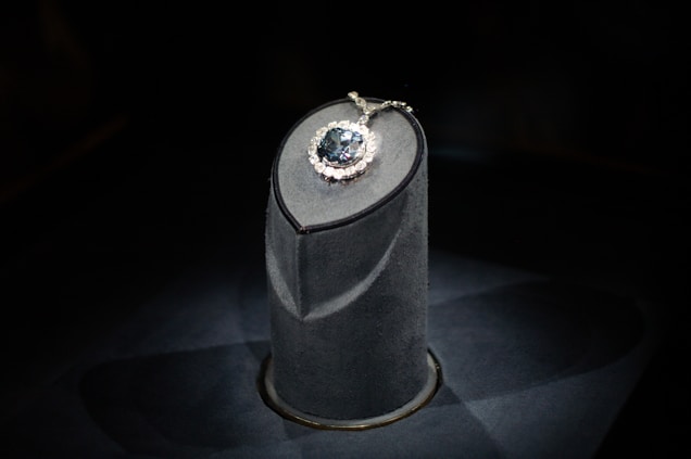 A luxurious gemstone necklace is displayed on a dark, elegant pedestal. The central gemstone is a deep, captivating blue, surrounded by smaller sparkling diamonds that enhance its brilliance. The spotlight enhances the sparkling effect, drawing attention to the intricate details and design.