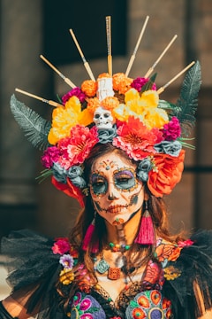 A person is adorned with vibrant Day of the Dead face paint, featuring intricate designs around the eyes and mouth. The individual wears an elaborate headdress with yellow, orange, pink, and red flowers, feathers, and a small skull at the top. The attire includes colorful embroidery and bright accessories.