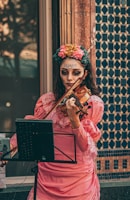 A woman wearing elaborate face makeup and a floral headpiece plays a violin in a public setting. She is dressed in a pink, vintage-style dress with lace detail, standing next to a tiled wall featuring geometric patterns.