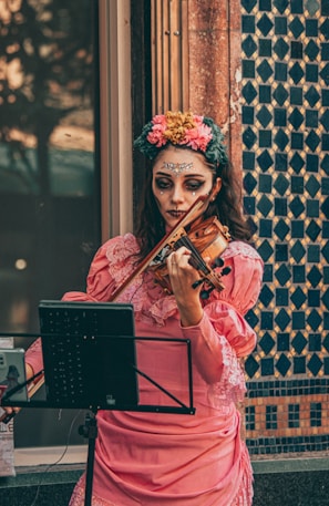 A woman wearing elaborate face makeup and a floral headpiece plays a violin in a public setting. She is dressed in a pink, vintage-style dress with lace detail, standing next to a tiled wall featuring geometric patterns.