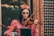 A woman wearing intricate face paint resembling a traditional Dia de los Muertos design with vibrant flowers in her hair plays a violin. She is dressed in a vintage pink dress with lace details, standing in front of a textured wall featuring geometric patterns.