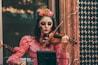 A woman wearing intricate face paint resembling a traditional Dia de los Muertos design with vibrant flowers in her hair plays a violin. She is dressed in a vintage pink dress with lace details, standing in front of a textured wall featuring geometric patterns.