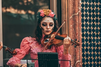 A woman wearing intricate face paint resembling a traditional Dia de los Muertos design with vibrant flowers in her hair plays a violin. She is dressed in a vintage pink dress with lace details, standing in front of a textured wall featuring geometric patterns.