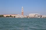 A picturesque waterfront scene featuring historic Italian architecture, with a prominent tall bell tower, ornate white buildings, and a vast body of water under a clear blue sky.
