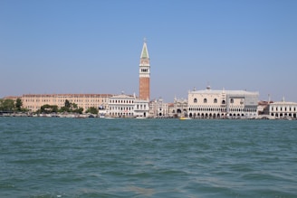 A picturesque waterfront scene featuring historic Italian architecture, with a prominent tall bell tower, ornate white buildings, and a vast body of water under a clear blue sky.