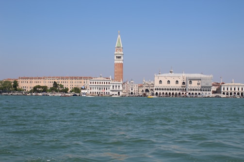 A picturesque waterfront scene featuring historic Italian architecture, with a prominent tall bell tower, ornate white buildings, and a vast body of water under a clear blue sky.