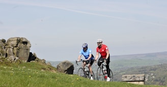 Two cyclists are riding on a grassy hillside with rocky outcrops in the background. One cyclist is wearing a blue jersey, and the other is in a red jersey, both equipped with helmets and riding road bikes. The background features a scenic countryside landscape with a partial view of a stone building and distant hills.