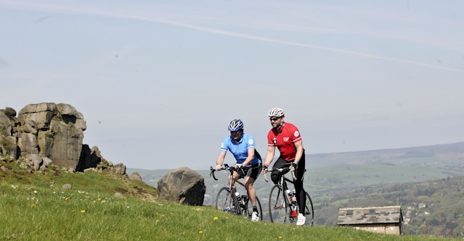 Two cyclists are riding on a grassy hillside with rocky outcrops in the background. One cyclist is wearing a blue jersey, and the other is in a red jersey, both equipped with helmets and riding road bikes. The background features a scenic countryside landscape with a partial view of a stone building and distant hills.