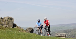 Two cyclists are riding on a grassy hillside with rocky outcrops in the background. One cyclist is wearing a blue jersey, and the other is in a red jersey, both equipped with helmets and riding road bikes. The background features a scenic countryside landscape with a partial view of a stone building and distant hills.