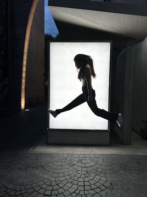 A silhouette of a person mid-air in front of a backlit panel, creating a striking contrast. The scene is set in an urban environment with textured pavement and an overhead structure partially visible.