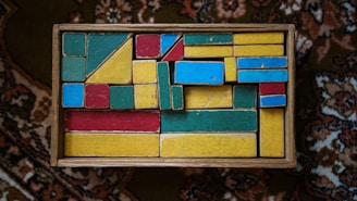 A box filled with variously shaped and colored wooden blocks, including rectangles, squares, and triangles in red, blue, yellow, and green. The arrangement of the blocks creates an intricate, geometric pattern. The box rests on a richly patterned carpet.