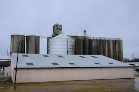 A large industrial complex featuring several tall, cylindrical silos and a smaller, rectangular building with a metal roof dominates the skyline. The silos are arranged in a row, some appearing metallic and others concrete, with various pipes and support structures connected to them. The overcast sky suggests gloomy weather and adds to the industrial feel of the scene.