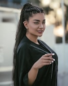 A woman with long, dark hair styled in braids and a ponytail, wearing a black outfit and decorative earrings. She has well-defined eyebrows, and her nails are manicured and painted white. The background is softly blurred, suggestive of an outdoor setting.