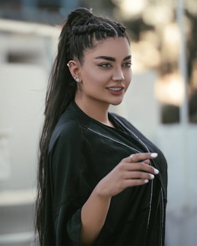 A woman with long, dark hair styled in braids and a ponytail, wearing a black outfit and decorative earrings. She has well-defined eyebrows, and her nails are manicured and painted white. The background is softly blurred, suggestive of an outdoor setting.