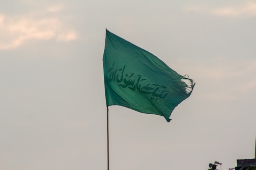 A green flag with Arabic script on it is flying against a cloudy sky. The flag appears slightly frayed at the edges and is attached to a pole.