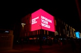 A brightly lit pink billboard with the text 'UPGRADE YOUR WORLD' is prominently displayed on the side of a building at night. The surrounding area is dark, but some other signs and lights are visible. The structure features vertical light patterns and a few street lamps, while Arabic script is seen on another part of the building.