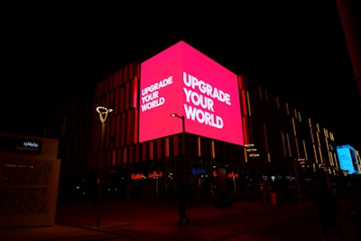 A brightly lit pink billboard with the text 'UPGRADE YOUR WORLD' is prominently displayed on the side of a building at night. The surrounding area is dark, but some other signs and lights are visible. The structure features vertical light patterns and a few street lamps, while Arabic script is seen on another part of the building.