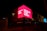 A brightly lit pink billboard with the text 'UPGRADE YOUR WORLD' is prominently displayed on the side of a building at night. The surrounding area is dark, but some other signs and lights are visible. The structure features vertical light patterns and a few street lamps, while Arabic script is seen on another part of the building.