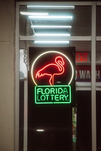 A neon sign depicting a flamingo above the words 'Florida Lottery' is prominently displayed. The sign is illuminated in bright red and green colors, and it is framed by glass panels. An 'EXIT' sign is visible in the background.