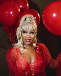 A woman is wearing a festive headband that says 'Birthday Queen' and a red lace outfit. She is surrounded by red balloons and has blonde hair styled in voluminous curls. The setting appears to be celebratory and vibrant.