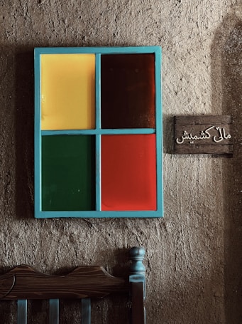 A rustic wall features a colorful, four-panel window with sections in yellow, brown, green, and red, surrounded by a light blue frame. To the right, a small wooden sign is mounted with Arabic script. Below the window, part of a wooden chair with a simple, sturdy design is visible.