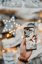A hand holding a smartphone displays a cozy scene of star-shaped string lights and soft fabrics. In the background, warm lights and a large decorative star create a festive ambiance.