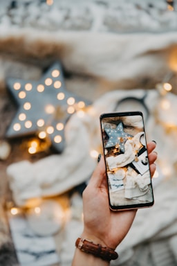 A hand holding a smartphone displays a cozy scene of star-shaped string lights and soft fabrics. In the background, warm lights and a large decorative star create a festive ambiance.