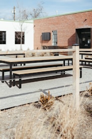 An outdoor space with wooden picnic tables and benches is situated on a gravel surface. The background features a brick building with a black door labeled 'Entrance' and a white building with several windows. Sparse dry grass and bushes are present across the area, adding to a barren landscape appearance. A wooden fence runs along the perimeter of the seating area.