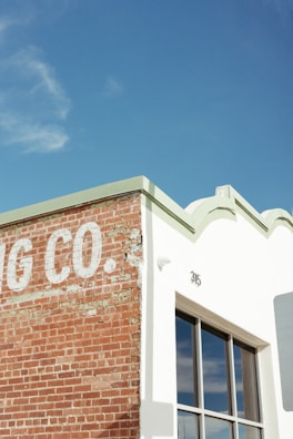 A corner of a building with a partially visible sign that reads 'G CO.' on a weathered brick wall, next to a white section with a large window reflecting the sky. The rooftop features decorative edges and the number 315 is visible.