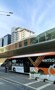 An electric bus in a modern urban environment is parked at a bus stop. The bus is orange and white with bold graphics and the text 'ELECTRIC BUS' on its side. There are glass buildings and a pedestrian in the background, alongside a multi-story building with a futuristic design.