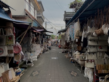 A narrow street lined with small shops displaying a variety of handcrafted items. These include dream catchers, woven bags, straw hats, and other artisanal products. The stalls are covered, and both sides of the street are densely packed with merchandise hanging and stacked.
