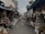 A narrow street lined with small shops displaying a variety of handcrafted items. These include dream catchers, woven bags, straw hats, and other artisanal products. The stalls are covered, and both sides of the street are densely packed with merchandise hanging and stacked.