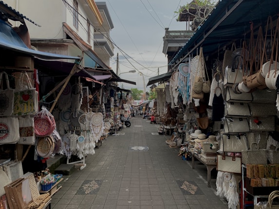 A narrow street lined with small shops displaying a variety of handcrafted items. These include dream catchers, woven bags, straw hats, and other artisanal products. The stalls are covered, and both sides of the street are densely packed with merchandise hanging and stacked.
