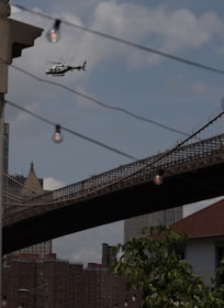A helicopter is flying in the sky near an urban area with a prominent suspension bridge in the background. The view includes buildings with a mix of architectural styles, and a few hanging light bulbs are visible in the foreground. The scene conveys a busy city environment.