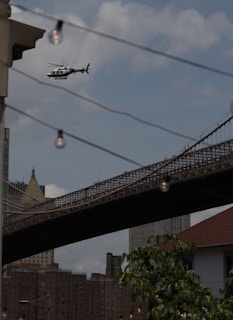 A helicopter is flying in the sky near an urban area with a prominent suspension bridge in the background. The view includes buildings with a mix of architectural styles, and a few hanging light bulbs are visible in the foreground. The scene conveys a busy city environment.