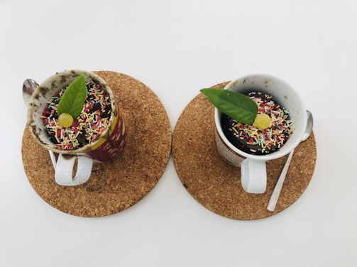 Two mugs of dessert topped with colorful sprinkles, green leaves, and small yellow decorations are placed on cork coasters. Each mug has a spoon beside it, and the background is a plain white surface.
