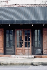 A storefront with a dark fabric awning over a pair of wooden double doors framed by two large glass windows. The building has a weathered appearance with visible bricks at the top and metal railings leading up to the entrance. A single black wire chair is placed on the right side near the entrance.