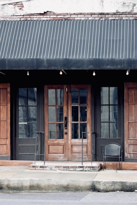 A storefront with a dark fabric awning over a pair of wooden double doors framed by two large glass windows. The building has a weathered appearance with visible bricks at the top and metal railings leading up to the entrance. A single black wire chair is placed on the right side near the entrance.