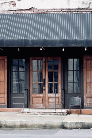 A storefront with a dark fabric awning over a pair of wooden double doors framed by two large glass windows. The building has a weathered appearance with visible bricks at the top and metal railings leading up to the entrance. A single black wire chair is placed on the right side near the entrance.