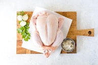 A raw whole chicken placed on a wooden cutting board, accompanied by fresh parsley, onions, and a small bowl of salt.