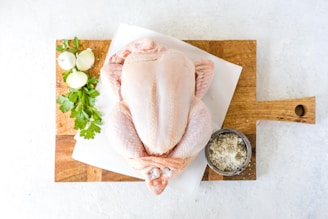 A raw whole chicken placed on a wooden cutting board, accompanied by fresh parsley, onions, and a small bowl of salt.