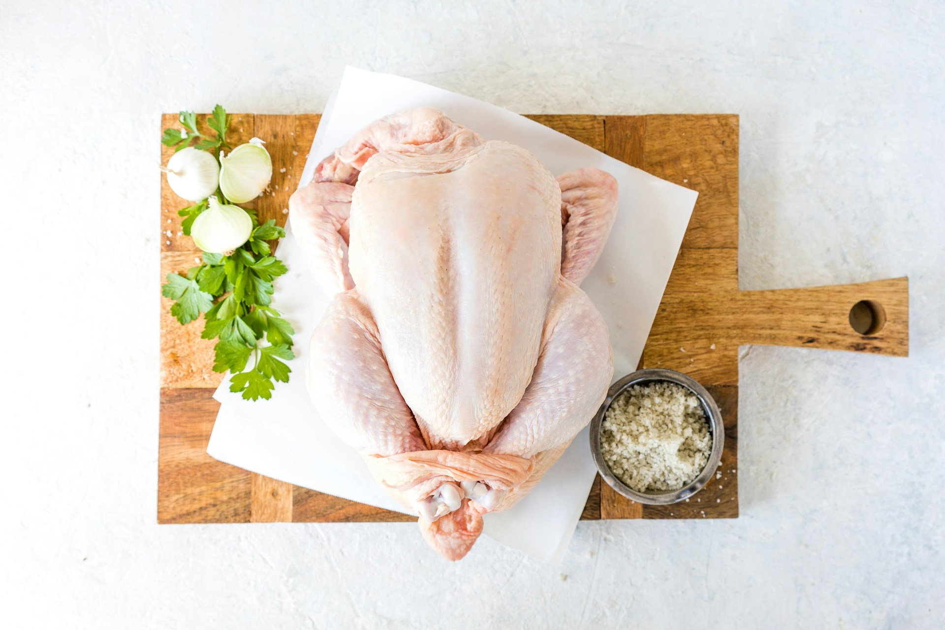 A raw whole chicken placed on a wooden cutting board, accompanied by fresh parsley, onions, and a small bowl of salt.