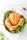 A perfectly roasted chicken is presented in a white dish surrounded by fresh herbs such as parsley and dill on a light-colored surface. The chicken has a golden brown, crispy skin, and there are visible specks of black pepper seasoning.