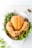 A perfectly roasted chicken is presented in a white dish surrounded by fresh herbs such as parsley and dill on a light-colored surface. The chicken has a golden brown, crispy skin, and there are visible specks of black pepper seasoning.