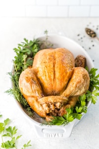 A perfectly roasted chicken is presented in a white dish surrounded by fresh herbs such as parsley and dill on a light-colored surface. The chicken has a golden brown, crispy skin, and there are visible specks of black pepper seasoning.