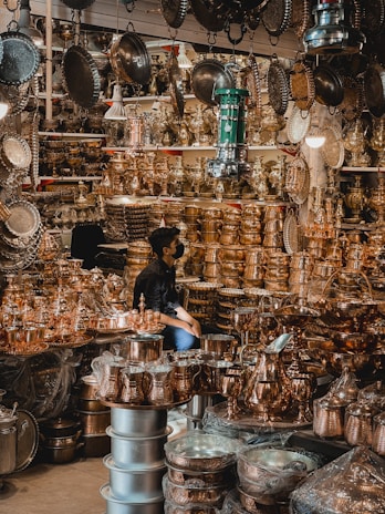 A busy market stall filled with a variety of shiny brass and copper kitchenware and decorative items. The items are stacked high on shelves and tables, reflecting the light and creating a warm, metallic ambiance. A person is sitting among the items, partially obscured by the abundance of metallic goods.